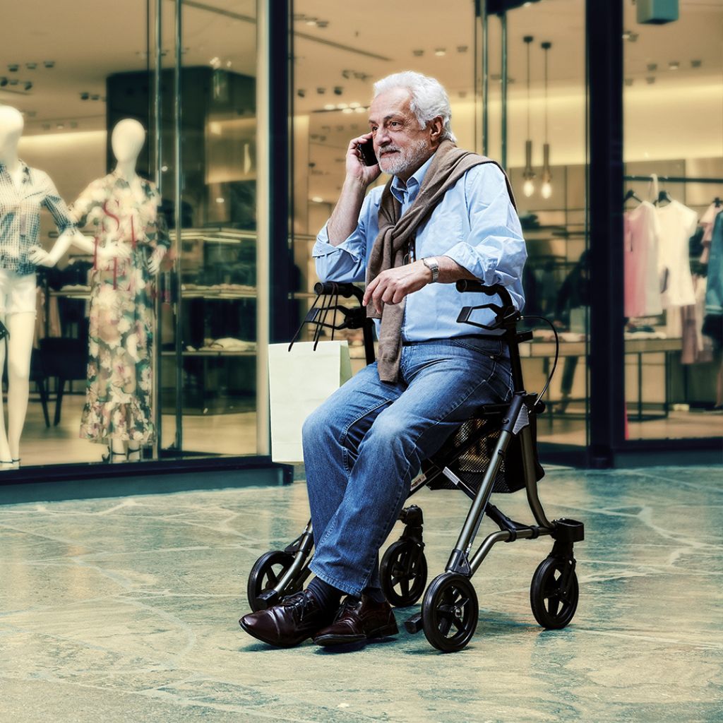 Dietz TAiMA M GT lightweight rollator in use in a shopping center, elderly man sitting on the rollator seat and talking on the phone.