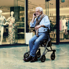 Dietz TAiMA M GT lightweight rollator in use in a shopping center, elderly man sitting on the rollator seat and talking on the phone.
