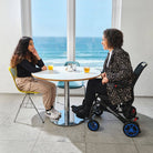 Zwei Frauen im Gespräch an einem Tisch mit Meerblick, eine davon sitzt in einem faltbaren Elektrorollstuhl.
